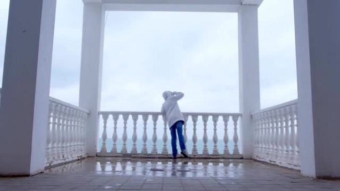 穿着白色夹克的女人在美丽的海景露台上欣赏大海。后视图。