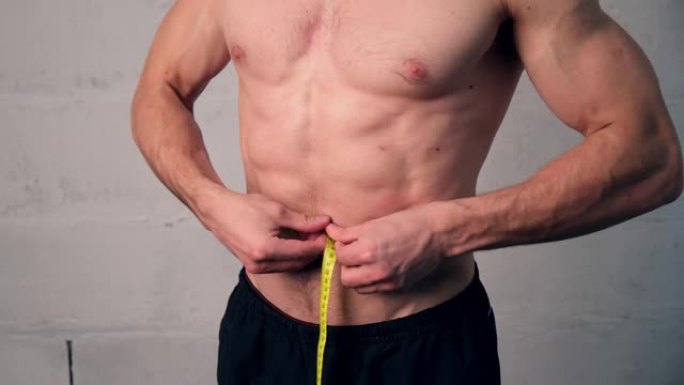 一个有着运动体格的年轻人测量他的腰围，检查他减掉了多少体重。减肥，男人过着运动的生活方式。一个健康的