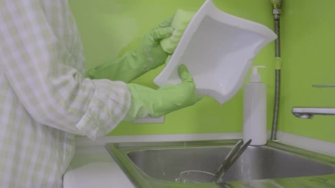 穿着橡胶手套的成熟女人在厨房用海绵和洗涤剂洗碗。