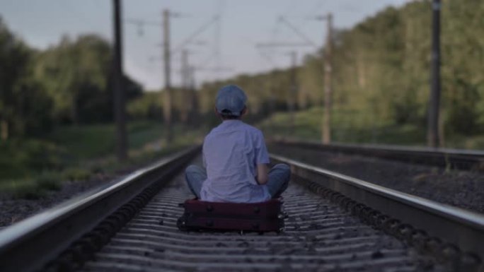 戴帽子的悲伤男孩坐在户外铁路上的手提箱上