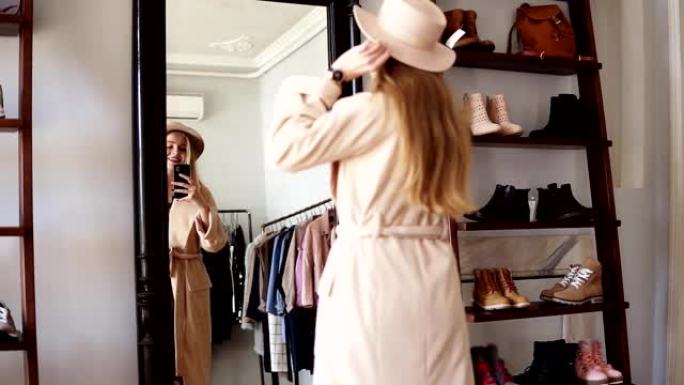 时尚的金发女孩站在镜子前，穿着米色外套和时髦的帽子，用智能手机拍了一张照片。享受她的风格。背面的镜头