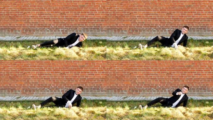一个穿着时髦的黑色西装的年轻高加索人躺在草地上，对着砖墙，慢吞吞的复制空间大笑