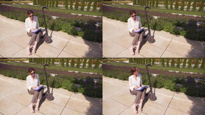 年轻的女主管坐在eco电动踏板车上的笔记本上制定了商业策略。城市景观中的成功、商业和生态交通概念