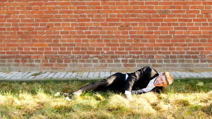 一个穿着黑色西装的年轻高加索人落在砖墙上的草地上，惨败，缓慢，复制空间