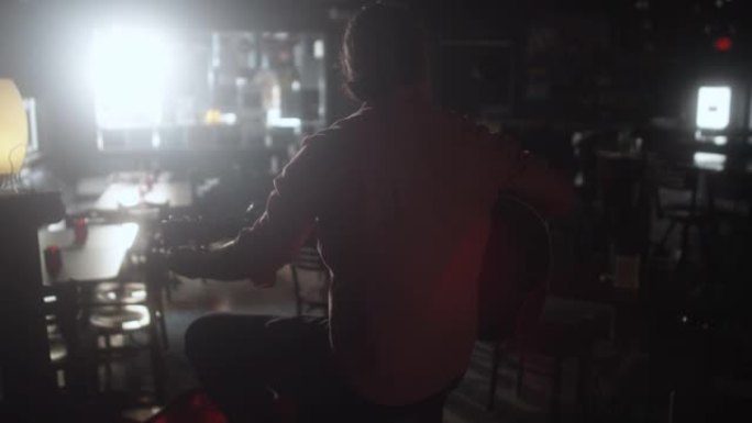 一个男人从后面在昏暗的酒吧里的一个小舞台上调音他的原声吉他，阳光普照