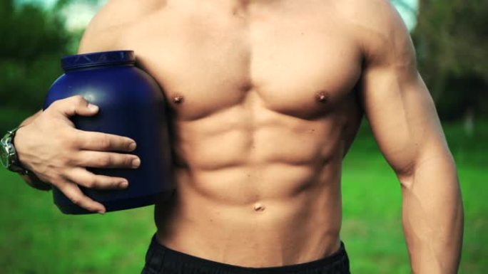 公园里肌肉发达的男性身体特写。职业运动员拿着一瓶营养补品。性感的健身教练示范运动食品添加剂的艰苦锻炼