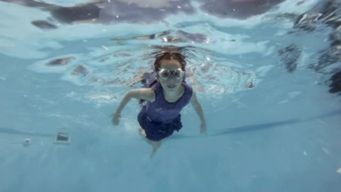 一个运动的小女孩戴着泳镜在游泳池水下游泳，看着相机。特写。底视图。慢动作。4K。