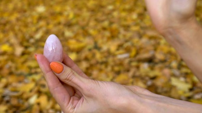橙色修指甲的女性手拿着粉色石英约尼蛋，适合秋季户外黄色落叶背景下的vumfit、imbuilding