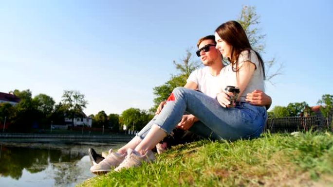 浪漫的情侣喝咖啡，笑着坐在池塘边的绿草地上。夫妻信任、爱和幸福的概念。完整镜头。