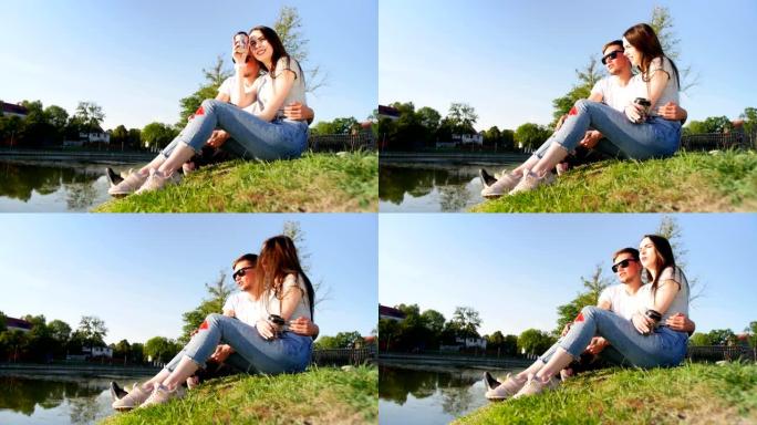 浪漫的情侣喝咖啡，笑着坐在池塘边的绿草地上。夫妻信任、爱和幸福的概念。完整镜头。