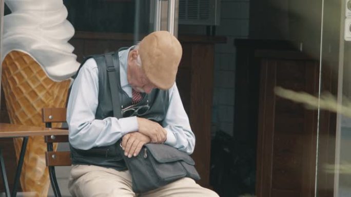 戴着灰色胡须和头发的帽子的老人坐在街上的椅子上睡着了。人们走过一个熟睡的老人。4k. 4k视频。60