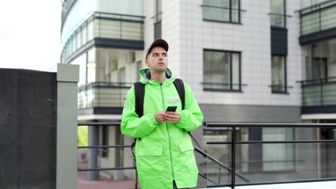 年轻的食品快递员穿着绿色制服，戴着隔热背包走在街上，上楼运送食物。在智能手机上使用移动导航应用程序的