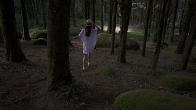 快乐的女孩旅行者带着草帽在魔法森林里奔跑，带着高大的绿色树木、植物和大神秘的石头。爱丽丝梦游仙境。