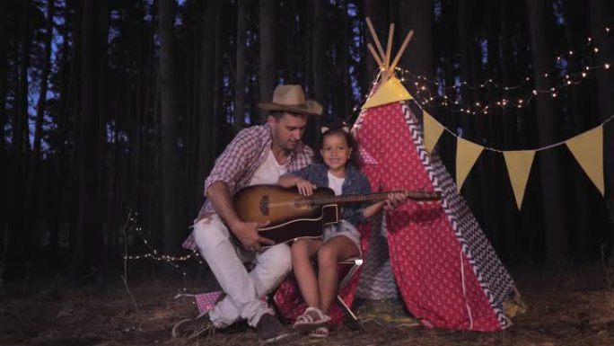 在森林里露营，快乐的父亲和可爱的女儿在户外度假时，在装饰着花环的棚屋边唱歌、弹吉他