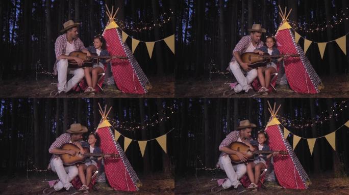 在森林里露营，快乐的父亲和可爱的女儿在户外度假时，在装饰着花环的棚屋边唱歌、弹吉他