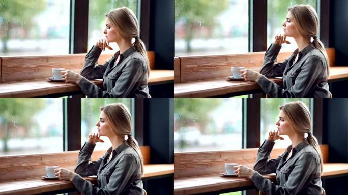 美丽沉思的金发不快乐的女人坐在咖啡馆里的雨天看着窗外