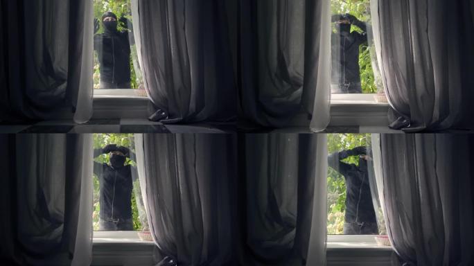 穿着黑色衣服和巴拉克拉法帽的小偷的特写镜头穿过窗户进入了房子。
