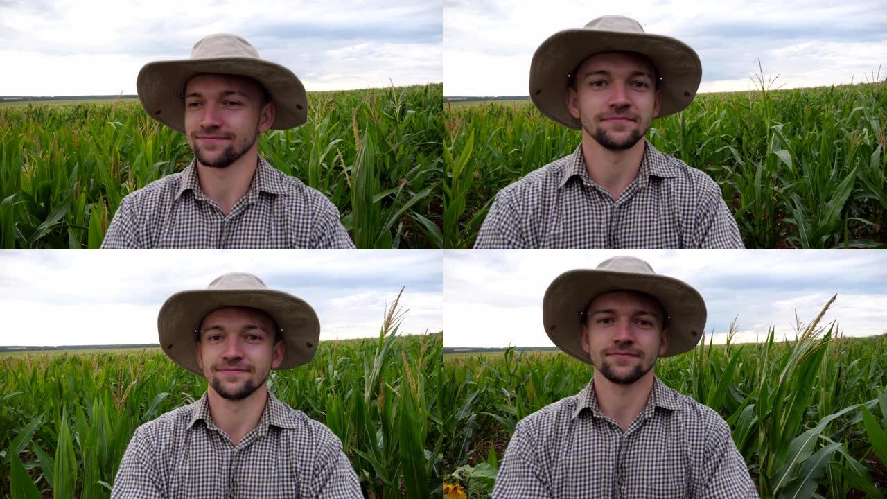 微笑工人在玉米种植园中自拍的男性脸的POV。戴着帽子的年轻快乐的农民在穿过玉米田时用智能手机射击自己