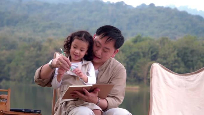 亚洲家庭父亲母亲和女儿在阳光明媚的夏天在公园的自然森林里露营和玩耍。父亲教写信。4k慢动作镜头。