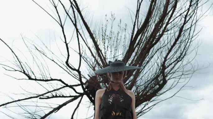 戴着黑帽子的漂亮金发女郎的肖像。女孩站在大干树下。