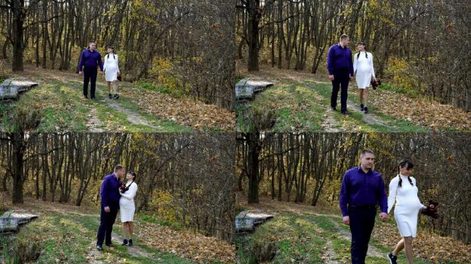一对怀孕的夫妇，一个年轻的女孩和一个男孩，在阳光明媚的日子里，牵着手在美丽的风景的背景下亲吻着秋天的