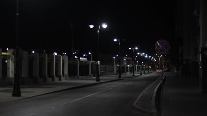 夏季可以看到夜间小巷，道路附近的交通标志，钢栅栏和城市路灯。库存镜头。没有人和汽车的安静夜街