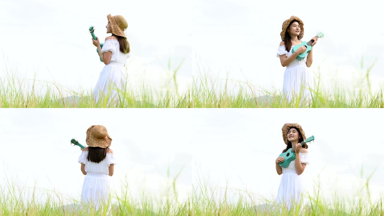 夏天，穿着白色连衣裙的亚洲幸福女人在绿色草地上玩尤克里里吉他，背景是山顶。音乐家美女用乐器放松。乡村