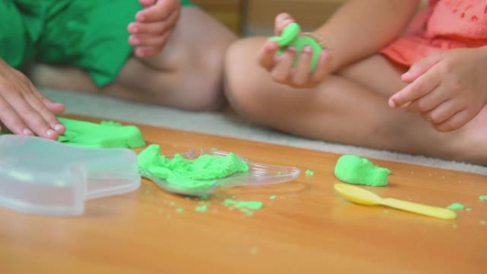 特写视频聚焦于小弟弟和妹妹、8岁男孩和6岁女孩的手，他们在住宅儿童房的地板上玩绿色的玩具沙子。照片与