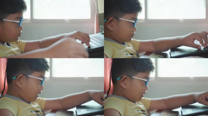 特写的快乐的亚洲儿童男孩戴着眼镜用笔记本电脑玩游戏，在一个房间里用智能手机看卡通，房间里有阳光从家里