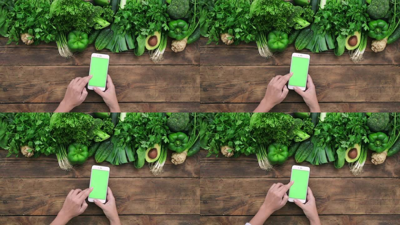 在线订购食物女人双手拿着手机，木质背景上有绿色模型，有干净的绿色蔬菜顶视图滚动手势做滑动
