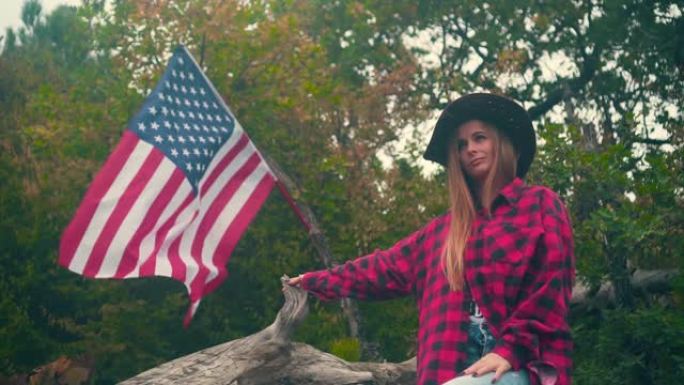 一个戴牛仔帽、穿格子衬衫的女孩站在一根圆木旁。背景是一面美国国旗在风中飘扬。