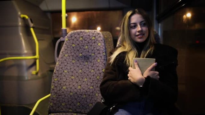 晚上乘公共汽车旅行时拿着书的女乘客。晚上，女孩在窗户旁边的行驶车辆上旅行