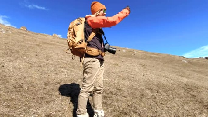一位快乐的女孩旅行者摄影师的肖像，戴着帽子和墨镜，脖子上戴着相机，手里拿着手机，在山峦的背景上拍照。