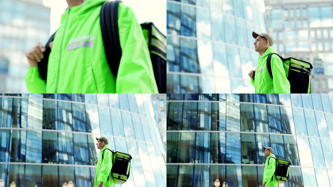 缩小跟踪左照片的年轻食品快递员穿着绿色制服，戴着隔热背包沿着城市街道行走。男子路过现代玻璃建筑交付订