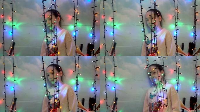 一个小女孩在房子的墙壁附近站在他们身后玩圣诞灯。她微笑着透过闪烁的灯光看着相机。特写。4K。