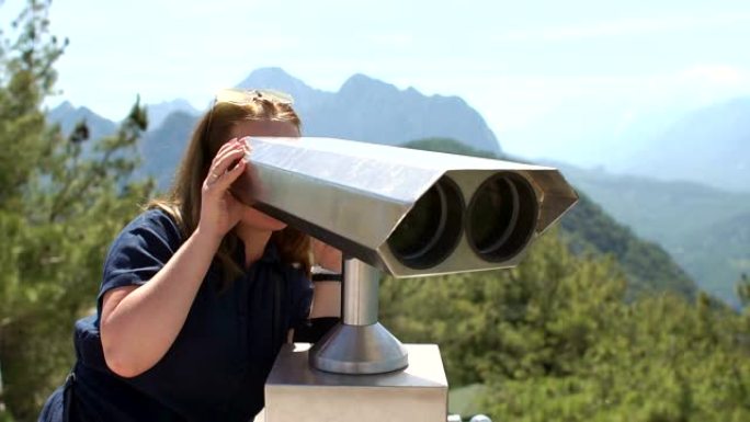 美丽的女孩正在山顶上看着投币双筒望远镜。