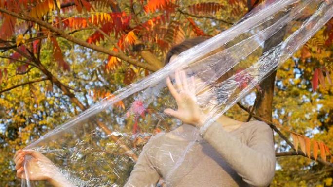 金发短发的年轻女子正透过塑料袋，聚乙烯包裹站在户外，在阳光明媚的秋天，靠近黄色和红色的叶子。