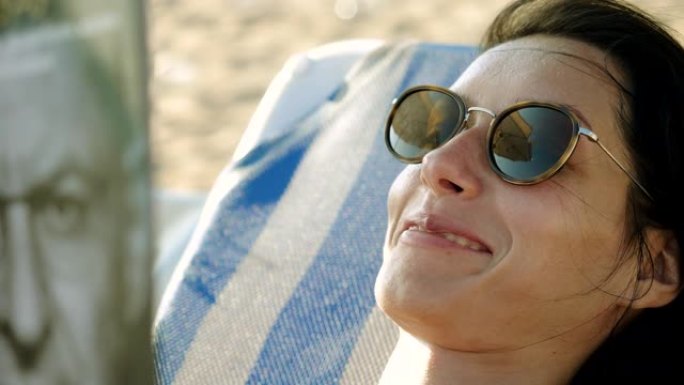 科斯塔德尔马雷斯梅海岸。美丽的年轻女子在海滩上度过一天的特写镜头，躺在遮阳伞下的日光浴躺椅上看书。卡