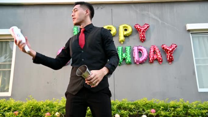 穿着黑色正装和红色领带的迷人英俊的亚洲男性酒保在兼职生日快乐活动中扮演杂耍演员。职业和人们生活方式娱
