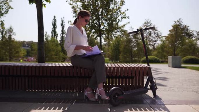 年轻独立的女商人坐在公园的长椅上，旁边是一辆电动踏板车。