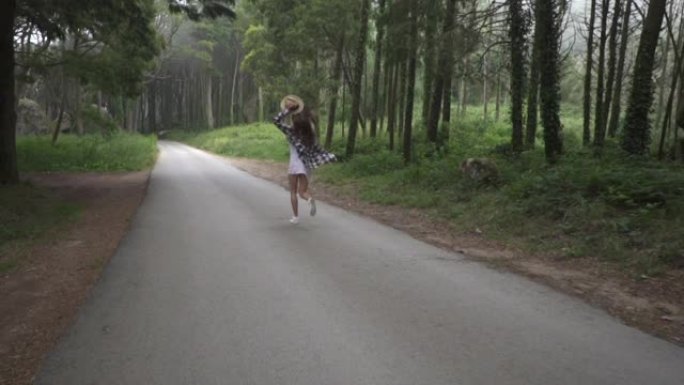美丽的旅行者女孩穿着衣服和草帽走在柏油路上，在神奇的神话般的森林里，有高树和大石头。辛特拉。葡萄牙。