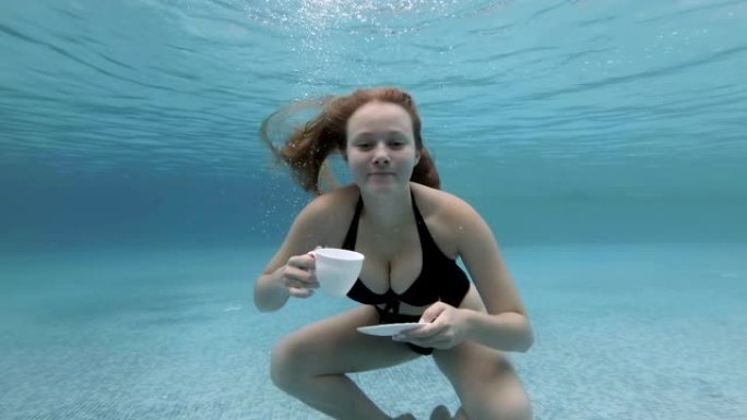 性感的年轻金发女郎长发潜水，穿着黑色泳衣，手里拿着白色杯子和碟子，在游泳池水下摆姿势。她吹泡泡，看着