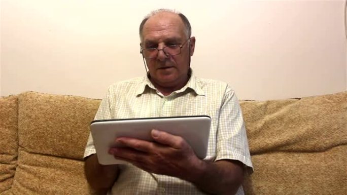 一位老人坐在家里的沙发上，在平板电脑上工作。
