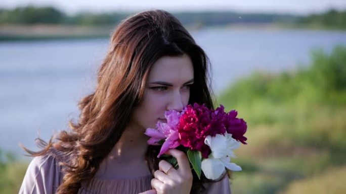 迷人的黑发女孩手里拿着一束鲜花。女孩将一束粉红色的牡丹压在胸前，嗅着花朵。