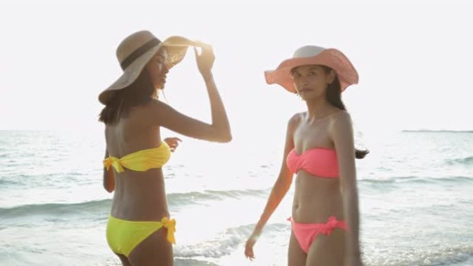两个穿着鲜艳比基尼的年轻女子在阳光明媚的早晨与大海漫步在沙滩上。