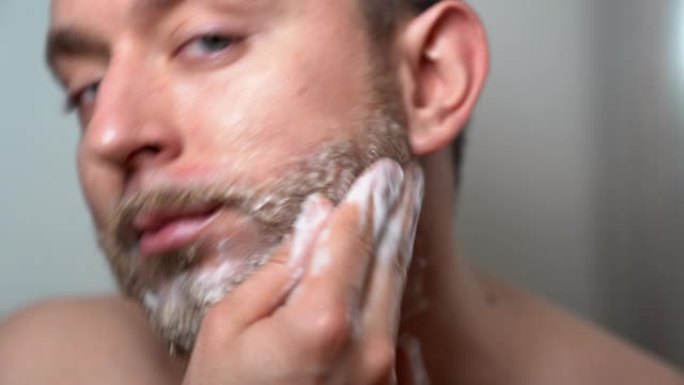 年轻英俊的男人用理发店的特制剃须洗发水给他未剃光的胡须擦肥皂。早上例行的野蛮男子在浴室，护肤和胡须卫