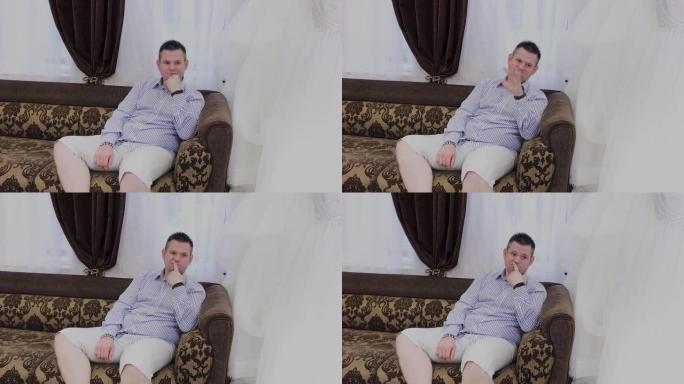 一个情绪激动的男人坐在婚礼沙龙的沙发上，对他的女人展示的衣服做出反应