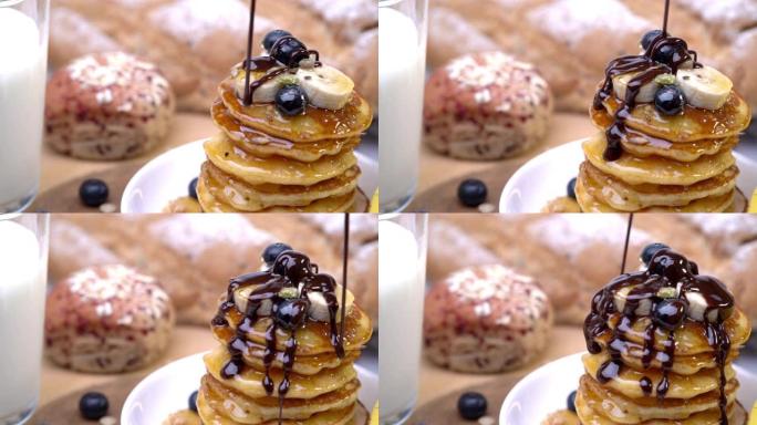 高清慢动作特写镜头将蜂蜜和巧克力糖浆倒在一堆自制的新鲜黄油煎饼上，上面放着蓝莓、全谷物和香蕉切片作为