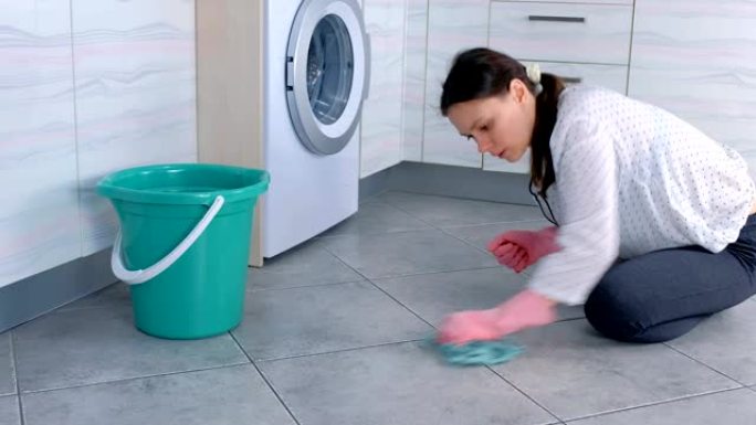 穿着粉红色橡胶手套的疲倦的女人用布擦洗和擦厨房地板上的污渍。地板上的灰色瓷砖。