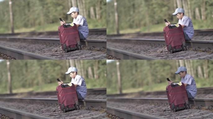 戴帽子的悲伤男孩坐在户外铁路上的手提箱上